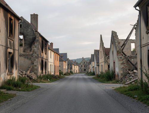 Vue respectueuse des ruines d’Oradour-sur-Glane au matin, avec une rue vide, des façades brûlées et une végétation discrète sous une lumière douce.