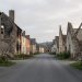Vue respectueuse des ruines d’Oradour-sur-Glane au matin, avec une rue vide, des façades brûlées et une végétation discrète sous une lumière douce.
