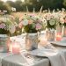 Table de fête champêtre avec des boîtes de conserve décorées transformées en vases, des photophores LED, des fleurs sauvages et des nappes en lin dans une douce lumière de fin d’après-midi.