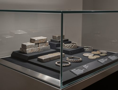 Vitrine muséale contemporaine présentant un trésor viking danois en argent composé de lingots, de bracelets et de monnaies anciennes, éclairé latéralement dans une ambiance archéologique élégante.