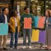 Groupe varié de femmes et d’hommes réunis devant une médiathèque de quartier, près d’affiches colorées vierges, dans une ambiance chaleureuse et réaliste.
